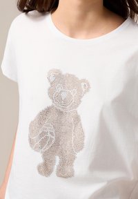 Weißes Baumwoll-T-Shirt mit einem Bärendesign, das aus kleinen, metallischen Strasssteinen besteht. Der Bär hält einen Basketball und hat eine strukturierten Optik.