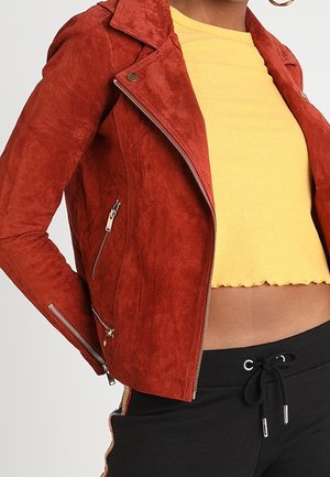 Kobieta w zamszowej kurtce w kolorze rdzawego pomarańczu, na żółtym skróconym prążkowanym topie oraz czarnych spodniach z trokami i bocznymi paskami.