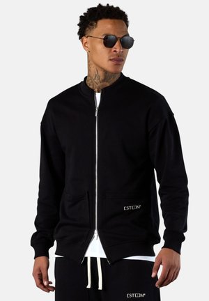 Man wearing black zip-up jacket and matching black pants with white drawstrings, sunglasses, and tattoos on his neck, standing against white background.