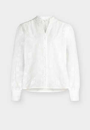 Biała bluzka z długim rękawem z subtelnym kwiatowym wzorem w żakardzie, zapinana na guziki z przodu, z marszczonymi mankietami na jednolitym tle.