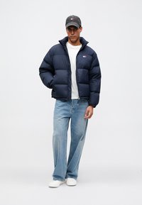 Άνδρας που φοράει μπορντό φούτερ μπουφάν, λευκή μπλούζα, ανοιχτό μπλε τζιν, λευκά αθλητικά παπούτσια και σκούρο καπέλο του μπέιζμπολ, στέκεται μπροστά σε απλό φόντο.