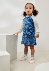Robe à imprimé léopard bleu avec manches grises, détail poche, et jupe à volants. Baskets blanches avec lanières pastel et chaussettes bleues. Fond neutre.