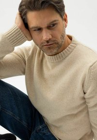 Mann mit braunen Haaren und Bart, der einen beigen Pullover und blaue Jeans trägt, sitzt mit einer Hand, die auf seinem Kopf ruht, und schaut nachdenklich nach unten.