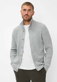 Graue Strickjacke mit Zopfmuster, mit hohem Kragen, Knopfleiste und strukturierten Ärmeln. Über einem weißen Hemd getragen.