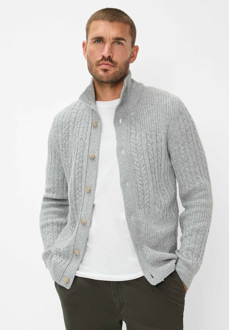 Graue Strickjacke mit Zopfmuster, mit hohem Kragen, Knopfleiste und strukturierten Ärmeln. Über einem weißen Hemd getragen.