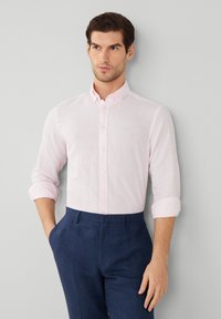 Lichtroze button-up shirt met strepen, lange mouwen tot de ellebogen opgerold, en een kleine geborduurde detail, gecombineerd met donkerblauwe trousers.