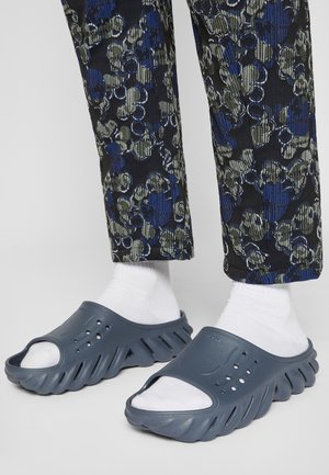 Sabots bleu foncé à bout ouvert avec une finition texturée et un design de semelle ondulée. Portés avec des chaussettes blanches et un pantalon à motifs verts et bleus.