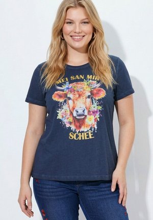 Lachende vrouw met blond haar draagt een marineblauw T-shirt met een kleurrijk koeienhoofd versierd met bloemen en de tekst "MEI SAN MIR SCHEE."