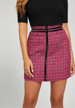 Kobieta nosząca różową mini spódnicę tweedową z czarnym pionowym suwakiem i czarną lamówką, zestawioną z czarną prążkowaną bluzką z krótkim rękawem.