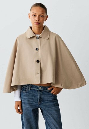 Mujer con una capa abotonada beige con cuello sobre una camisa blanca de manga larga y vaqueros azules, de pie con una mano en el bolsillo.