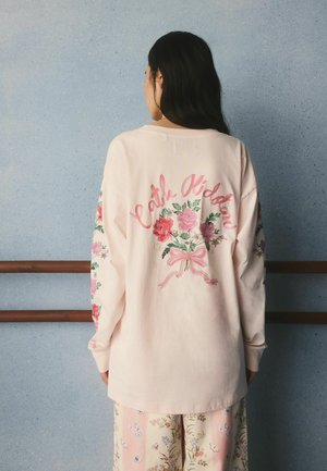 Frau mit langen dunklen Haaren, die ein hellrosa Langarmshirt mit Blumen- und Schleifendekor sowie dem Text "Cath Kidston" auf dem Rücken trägt.