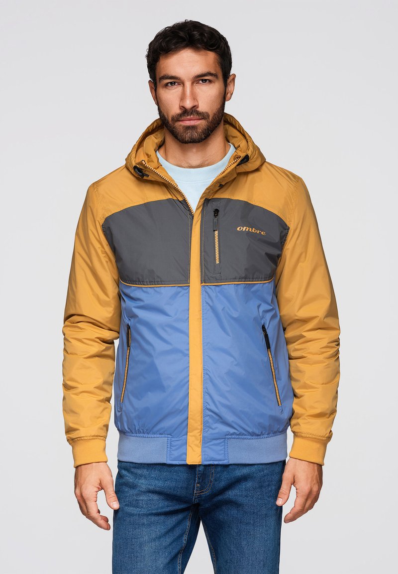 Homme portant une veste à capuche jaune moutarde, grise et bleue avec des poches zippées et un jean bleu, debout devant un fond clair uni.