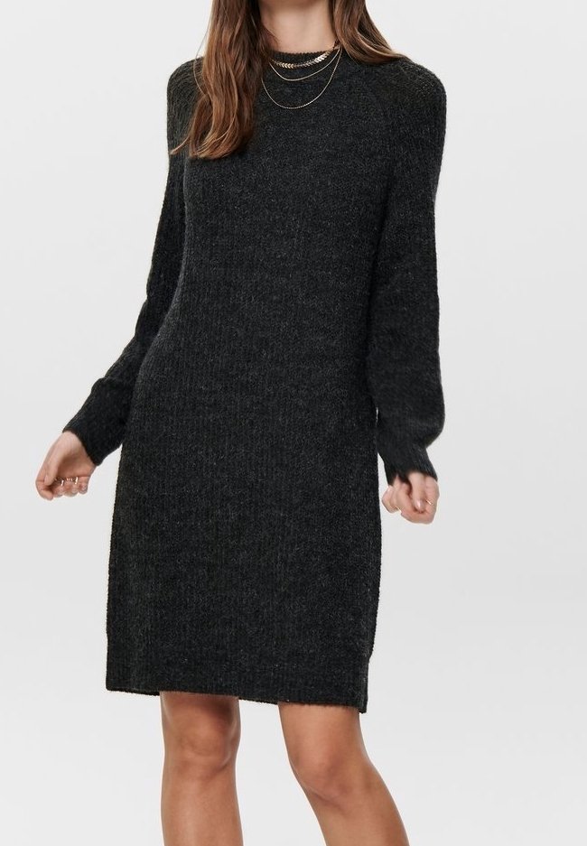 Femme portant une robe-pull en maille gris foncé à manches longues et des colliers en or superposés, debout devant un fond uni.