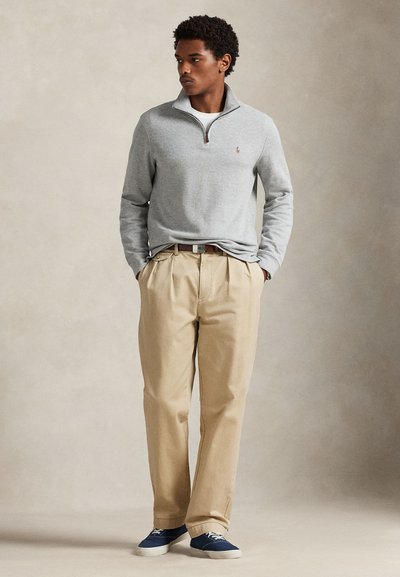 Homme debout les mains dans les poches portant un pull gris à demi-fermeture éclair, pantalon beige, ceinture marron et baskets bleues sur un fond neutre.