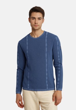 Junger Mann trägt einen strukturierten blauen Langarmpullover und beige Hose, steht vor einem schlichten weißen Hintergrund.