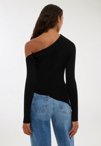 Kobieta z długimi ciemnymi włosami nosi czarny asymetryczny sweter bez ramion i niebieskie dżinsy, odwrócona przodem do białego tła.