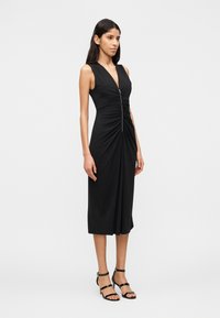 Γυναίκα με μαύρο midi φόρεμα χωρίς μανίκια, με φερμουάρ μπροστά και ρυθμιζόμενη λεπτομέρεια, συνδυασμένο με μαύρα σανδάλια με λουράκια και ψηλό τακούνι.