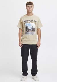 Beige grafisk t-skjorte med teksten "OUTDOORS" og et landskapsmotiv. Kombinert med svarte cargo-bukser og hvite sneakers. Uformelt gateklesantrekk.
