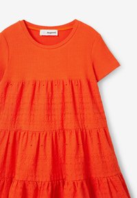 Oranžové šaty s krátkými rukávy. Žebrované živůtek a vícedílná sukně s detailním vyšíváním, které vytváří texturu a vizuální zájem.