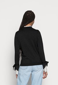 Pull noir avec une finition texturée, manches longues avec des liens aux poignets et un col rond. Porté avec un jean bleu clair.