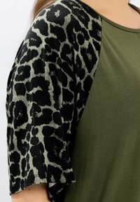 Close-up van een persoon die een olijfgroen shirt draagt met korte mouwen in zwart en donkergroen dierenprint.