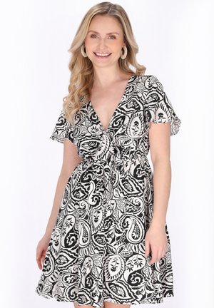 Donna sorridente con lunghi capelli biondi che indossa un vestito a fantasia floreale bianco e nero con scollo a V e maniche corte a svolazzo, in piedi davanti a uno sfondo bianco.