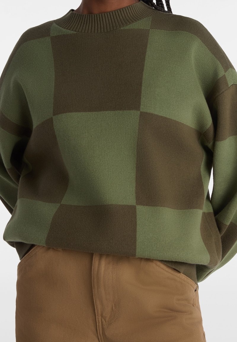 Grön rutigt stickad tröja med ribbat halsringning. Löst sittande, med kontrasterande rutor i mörk och ljusgrön. Kombinerad med beiga byxor.