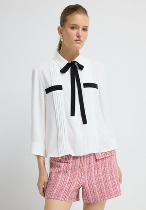 Hvid bluse med sorte accenter og en sløjfe, med vertikale folder og en afslappet pasform; parret med teksturerede lyserøde tweed shorts.