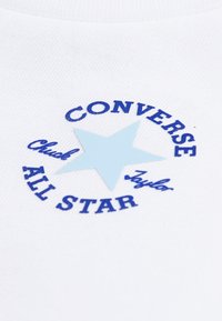 Λευκό βαμβακερό μπλουζάκι με μπλε κυκλικό λογότυπο που περιλαμβάνει ένα ανοιχτό μπλε αστέρι, κείμενο "CONVERSE ALL STAR" και τη φράση "Chuck Taylor" σε μπλε γραφή.