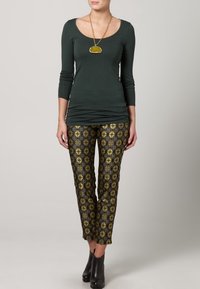 Tmavě zelený top s dlouhými rukávy a kulatým výstřihem, kombinovaný s černými a zlatými vzorovanými úzkými kalhotami a kotníkovými botami. Výrazný zlatý náhrdelník.