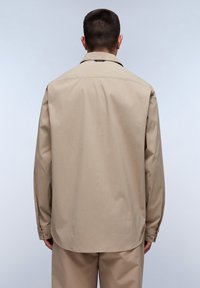 Camicia beige a maniche lunghe con colletto classico e polsini con bottoni. Realizzata in tessuto liscio, presenta una vestibilità leggermente ampia e un giogo sulla schiena.