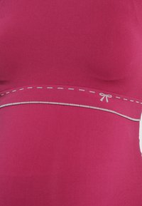 Fitted top in bold pink fabric featuring a decorative grey stitch along the hem and a small bow detail at the center. Smooth texture.