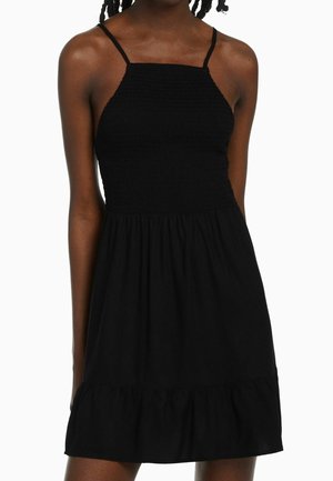 Vestido negro de tela ligera. Cuenta con un top fruncido, tirantes delgados y una falda acampanada con un dobladillo con volantes. Diseño simple y casual.