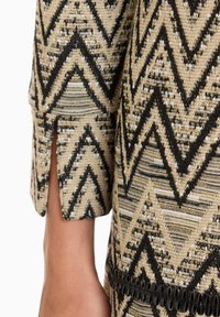 Gemustertes Kleidungsstück mit einem Chevron-Muster in Beige, Schwarz und Gold. Enthält einen seitlichen Schlitz und dekorative schwarze Einfassung am Saum.