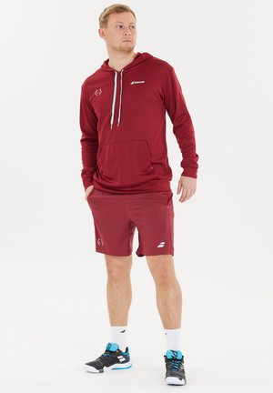 Bordeaux hættebluse med snoretræk, frontlomme; parret med matchende shorts. Fodtøjet inkluderer sorte sneakers med blå accenter.