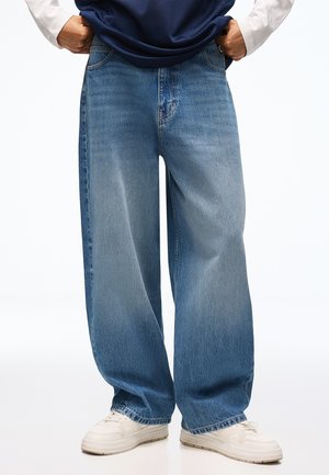 Person trägt weit geschnittene blaue Jeans und weiße Sneakers, die Hände in den Jeanstaschen, vor einem einfarbigen Hintergrund.