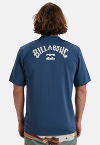 Camiseta de manga corta azul marino con un logo blanco "BILLABONG" y diseño de ola en la parte posterior. Textura suave, corte holgado.