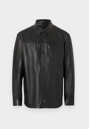 Μαύρο πουκάμισο-σακάκι από δερματίνη με μυτερό γιακά, κούμπωμα με κουμπιά και δύο κάθετες τσέπες στο στήθος με φερμουάρ, εμφανίζεται σε απλό φόντο.