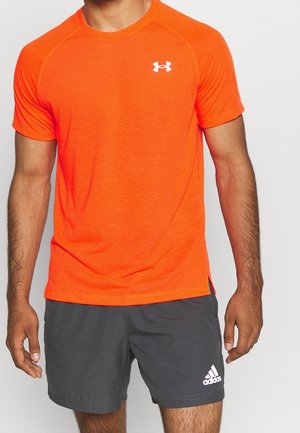 Mężczyzna w jaskrawo pomarańskiej koszulce z krótkim rękawem Under Armour i ciemnoszarych sportowych spodenkach Adidas, stoi z rozluźnionymi ramionami.