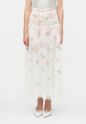 Mujer que lleva una falda blanca transparente con bordado floral rosa y tacones blancos puntiagudos con calcetines de encaje, de pie frente a un fondo liso.