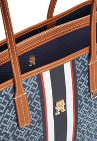 Τσάντα tote με εξωτερική επιφάνεια σε μπλε μοτίβο, καφέ δερμάτινη επένδυση, χρυσαφένια εξαρτήματα και κλείσιμο με φερμουάρ. Διαθέτει κόκκινες και λευκές ρίγες.