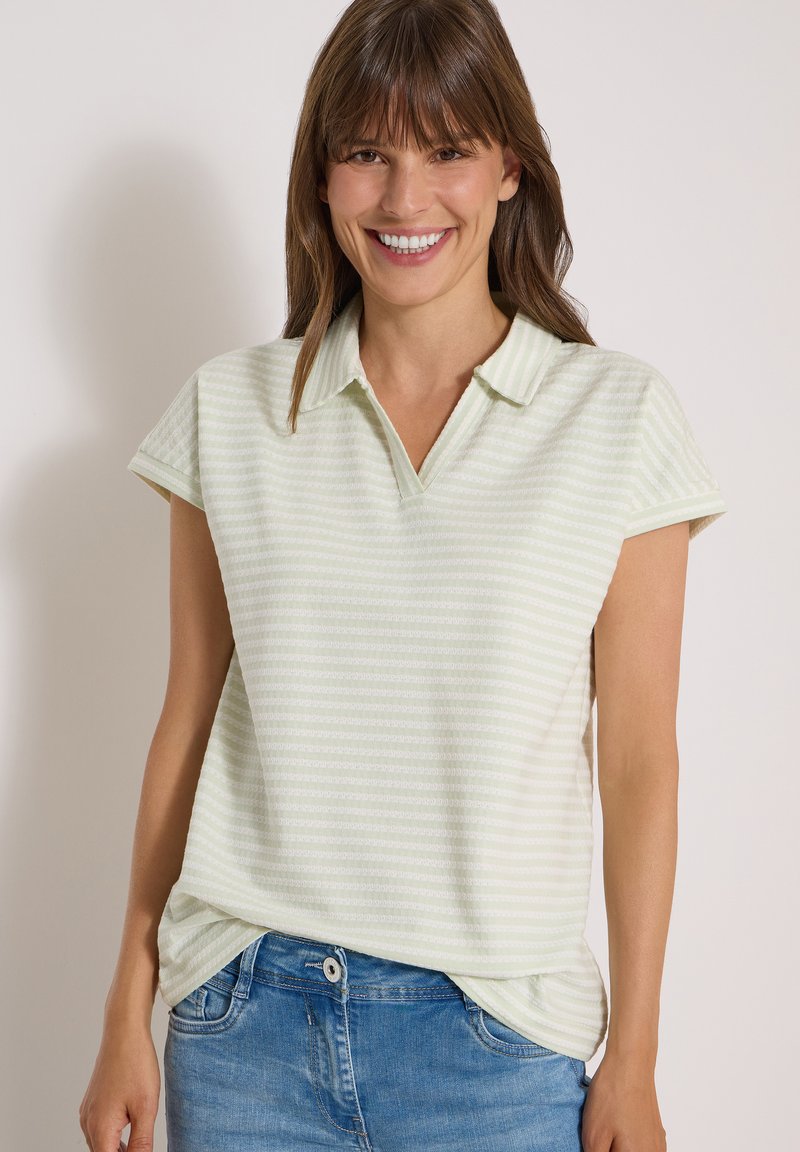 Femme souriante portant un polo à manches courtes rayé vert clair et un jean bleu sur fond uni.