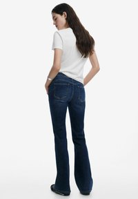 Jean en denim bleu foncé évasés avec une coupe à cinq poches, présentant des coutures contrastées et un détail de fermeture éclair à l'arrière. Associé à un t-shirt blanc.