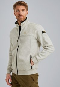 Veste en polaire gris clair avec un col haut, un zip à l'avant et des accents noirs ; elle possède une poche poitrine et des poches latérales.