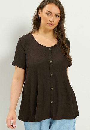 Femme portant un haut ample à manches courtes boutonné marron foncé, associé à un jean bleu clair sur un fond uni clair.