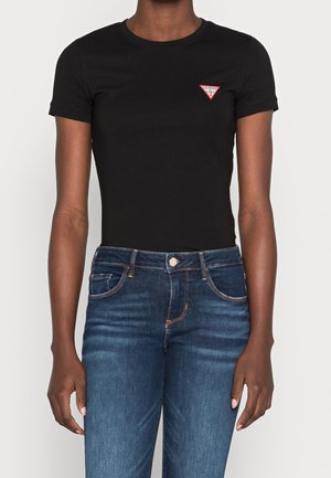 Person iført en tætsiddende sort t-shirt med et lille rødt og hvidt Guess-logo på brystet og blå denimjeans.