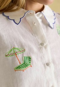 Weißes Leinenhemd mit einem gewellten Kragen und blauer Stickerei. Das bestickte Design zeigt einen grünen Regenschirm und einen gestreiften Liegestuhl.