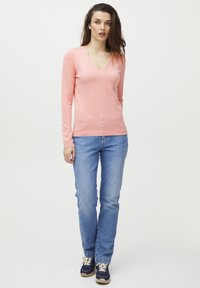 Un pull V en laine rose clair à manches longues, confectionné dans un matériau doux, associé à un jean droit bleu clair et des baskets bleues avec des lacets blancs.