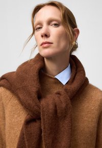 Écharpe en tricot marron enroulée autour d'un pull beige, avec une chemise à col bleu clair visible en dessous. Texture douce et design superposé.