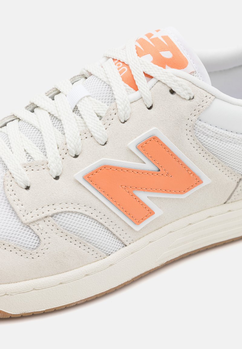 Baskets avec une tige en maille blanche et en daim beige, comportant un logo "N" orange bien en évidence. Semelle extérieure en caoutchouc texturé avec des coutures détaillées.